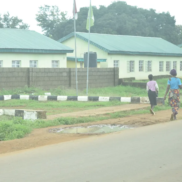A Nigerian secondary school campus: white classroom blocks, a school signboard and a paved road with kerbs. Openly licensed photograph by Kambai Akau.